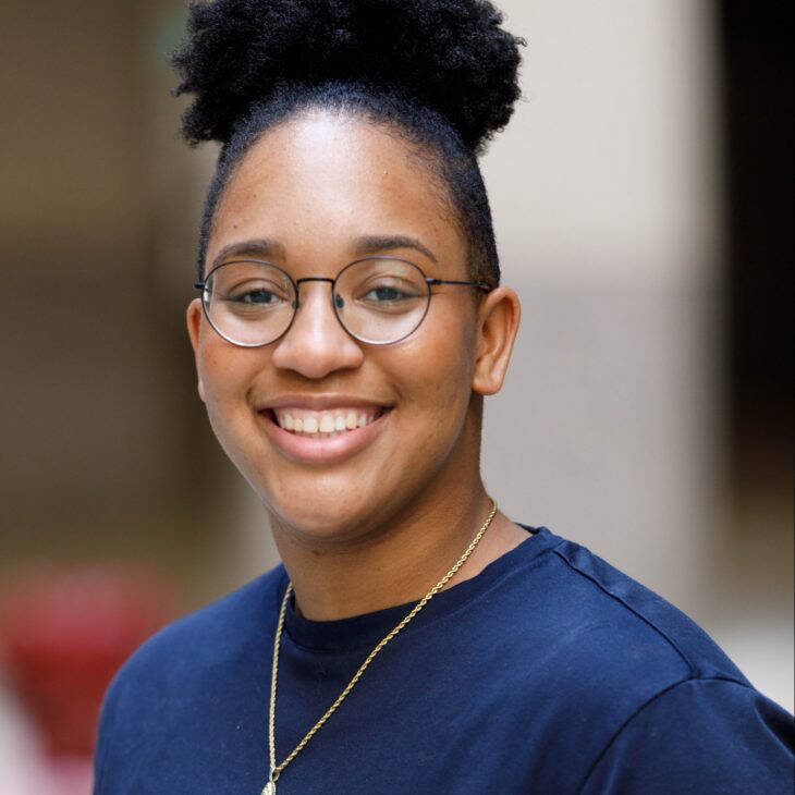 A black person in glasses and a blue t-shirt in front of a blurry background.
